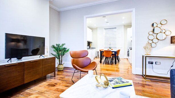 Una sala de estar moderna con un televisor de pantalla plana, un sillón de cuero marrón, una mesa de centro blanca y un comedor con sillas naranjas.