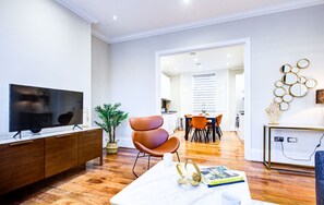 Ein modernes Wohnzimmer mit Flachbildfernseher, einem braunen Ledersessel, einem weißen Couchtisch und einem Essbereich mit orangefarbenen Stühlen.