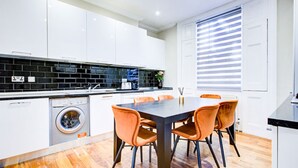 Una cocina moderna con armarios blancos, un salpicadero de azulejos negros, lavadora integrada y un comedor con mesa y sillas naranjas.