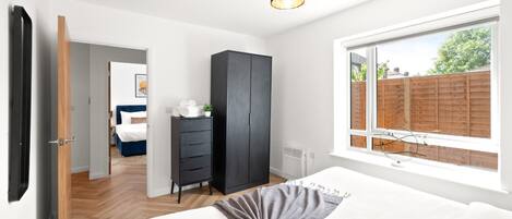 A bedroom with a bed, a wardrobe, a dresser, a window with a view of a wooden fence, and a door leading to another room.