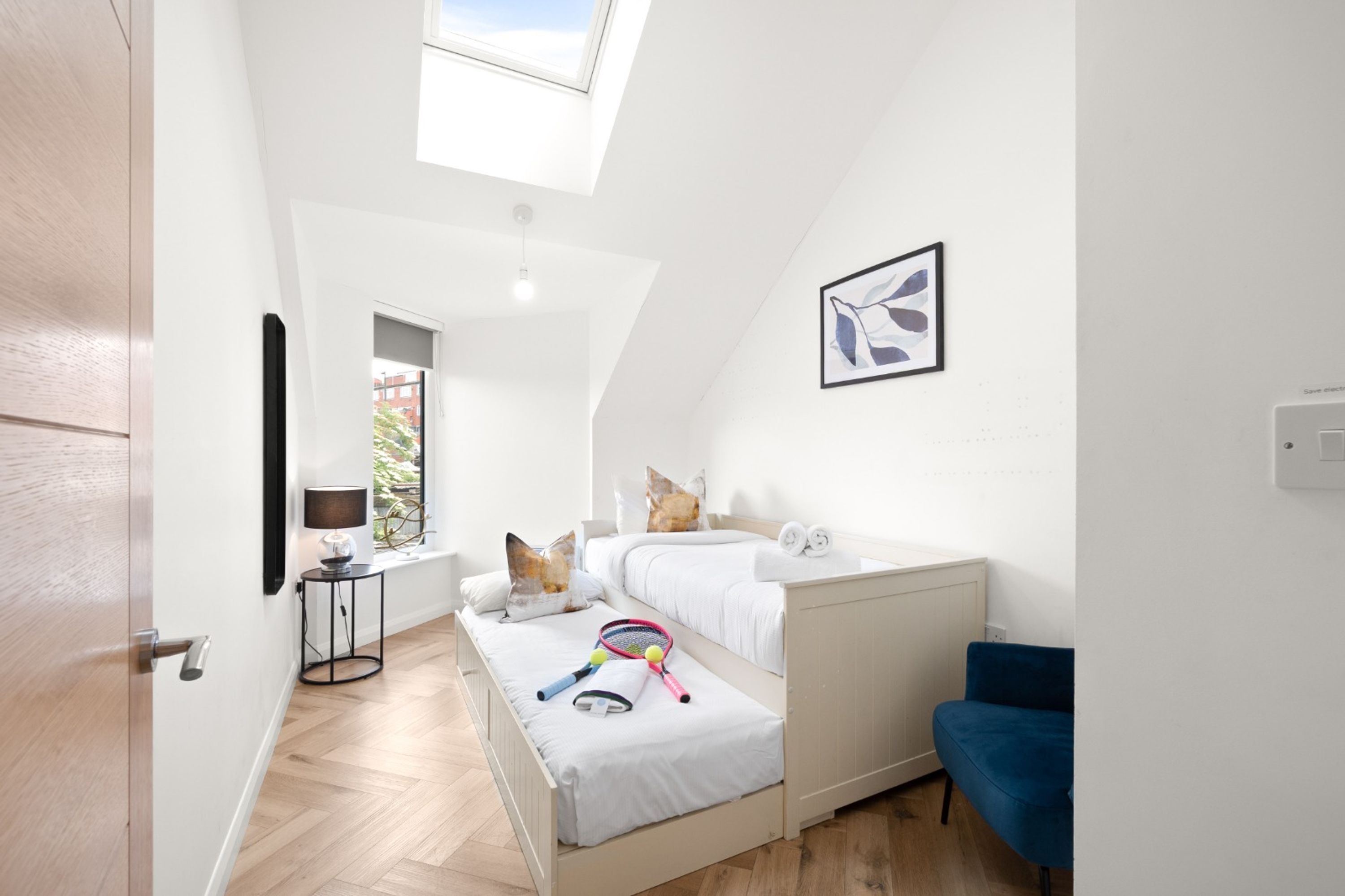 Ein Schlafzimmer mit schrägverdeckter Decke, einem Bett, einem Nachttisch mit Lampe, einem Fenster und einem an der Wand befestigten Lichtschalter.