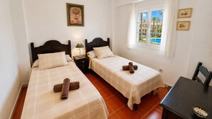 A hotel room with two beds, a nightstand with a lamp, a window with a view of a pool, and a ceiling fan. - Alcaraván T2, Sea view apartment - Ref. 11P21 (Ayamonte)