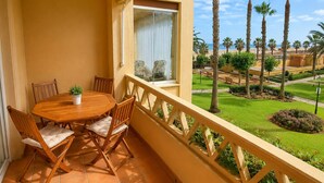 A balcony with a wooden table and chairs, a potted plant, and a view of a well-maintained garden with palm trees and a beach in the background. - Alcaraván T2, Sea view apartment - Ref. 11P21 (Ayamonte)