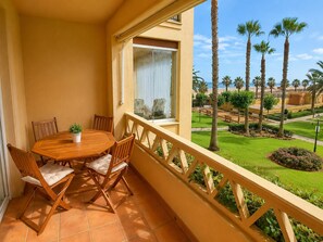 Ein Balkon mit Holztisch und -stühlen, einem Blumentopf und Blick auf einen gepflegten Garten mit Palmen und einen Strand im Hintergrund.