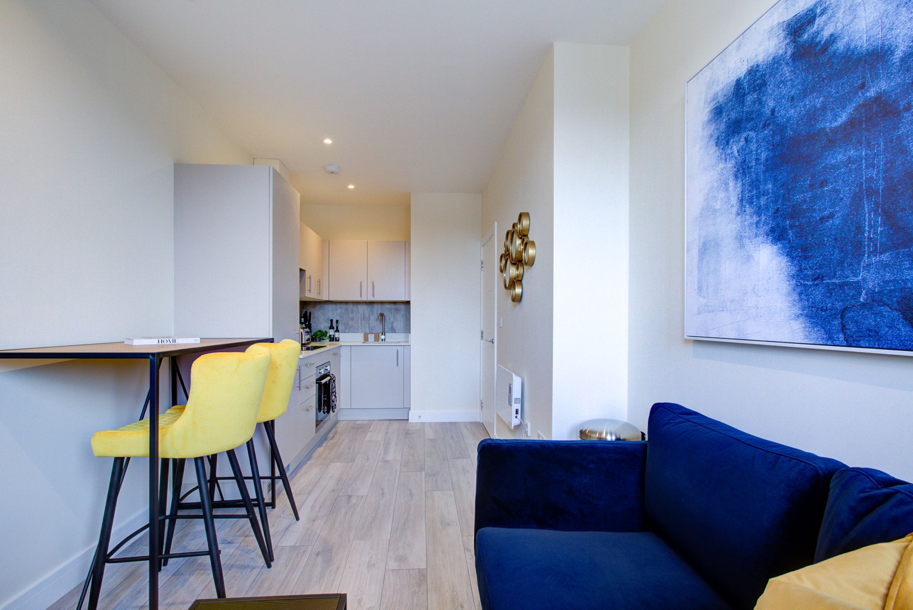 Ett kompakt bostadsutrymme med ett litet kök, en matplats med gula stolar och en blå soffa.