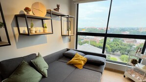 Una sala de estar moderna con un sofá, cojines, un reloj, estanterías con plantas y un amplio ventanal con vistas a la ciudad.