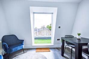 A room with a large window, a blue chair, a dining table with a plant, and a rug on the floor. - Hybrid Resi - Wandsworth - Apartment (London)