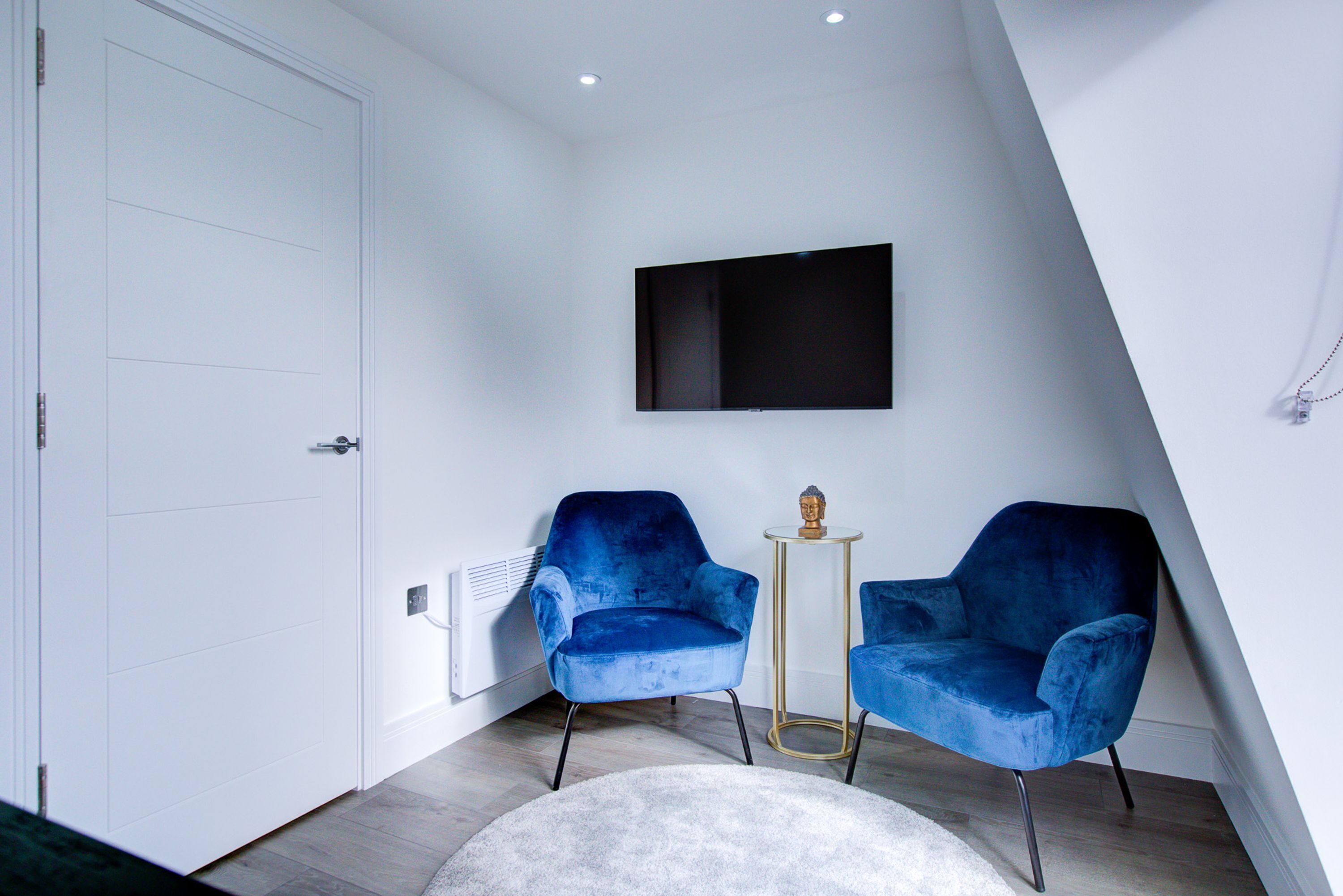 A modern room with two blue chairs, a small round table, a mounted flat-screen TV, and a white door.