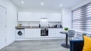 A modern kitchen with white cabinetry, a washing machine, an oven, and a dining area with a table and chairs. - Hybrid Resi - Wandsworth - Apartment (London)