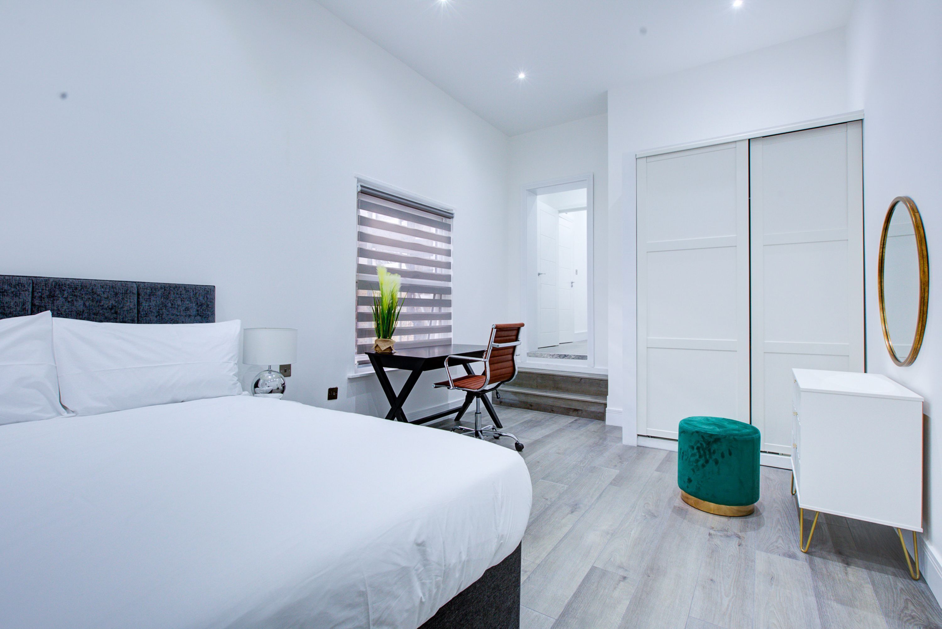 A modern hotel room with a large bed, a desk, a chair, a wardrobe, and a small ottoman.