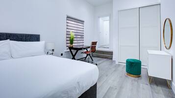 A modern hotel room with a large bed, a desk, a chair, a wardrobe, and a small ottoman.