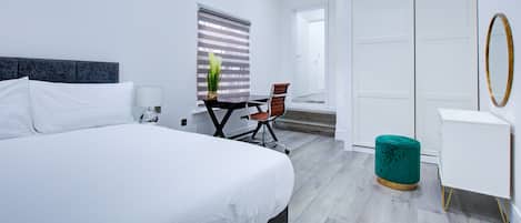 A modern hotel room with a large bed, a desk, a chair, a wardrobe, and a small ottoman.