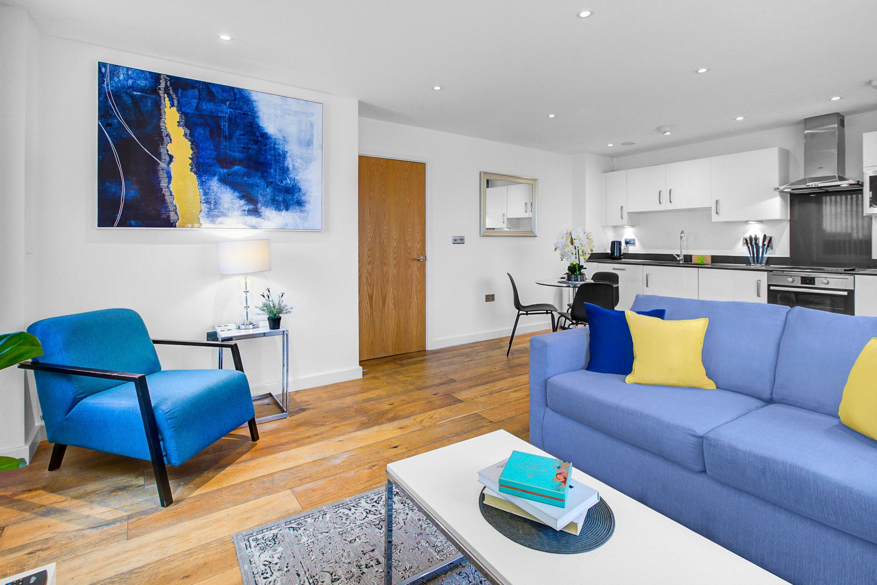 A modern living room with a blue sofa, a coffee table with books, a blue armchair, and a wooden floor.