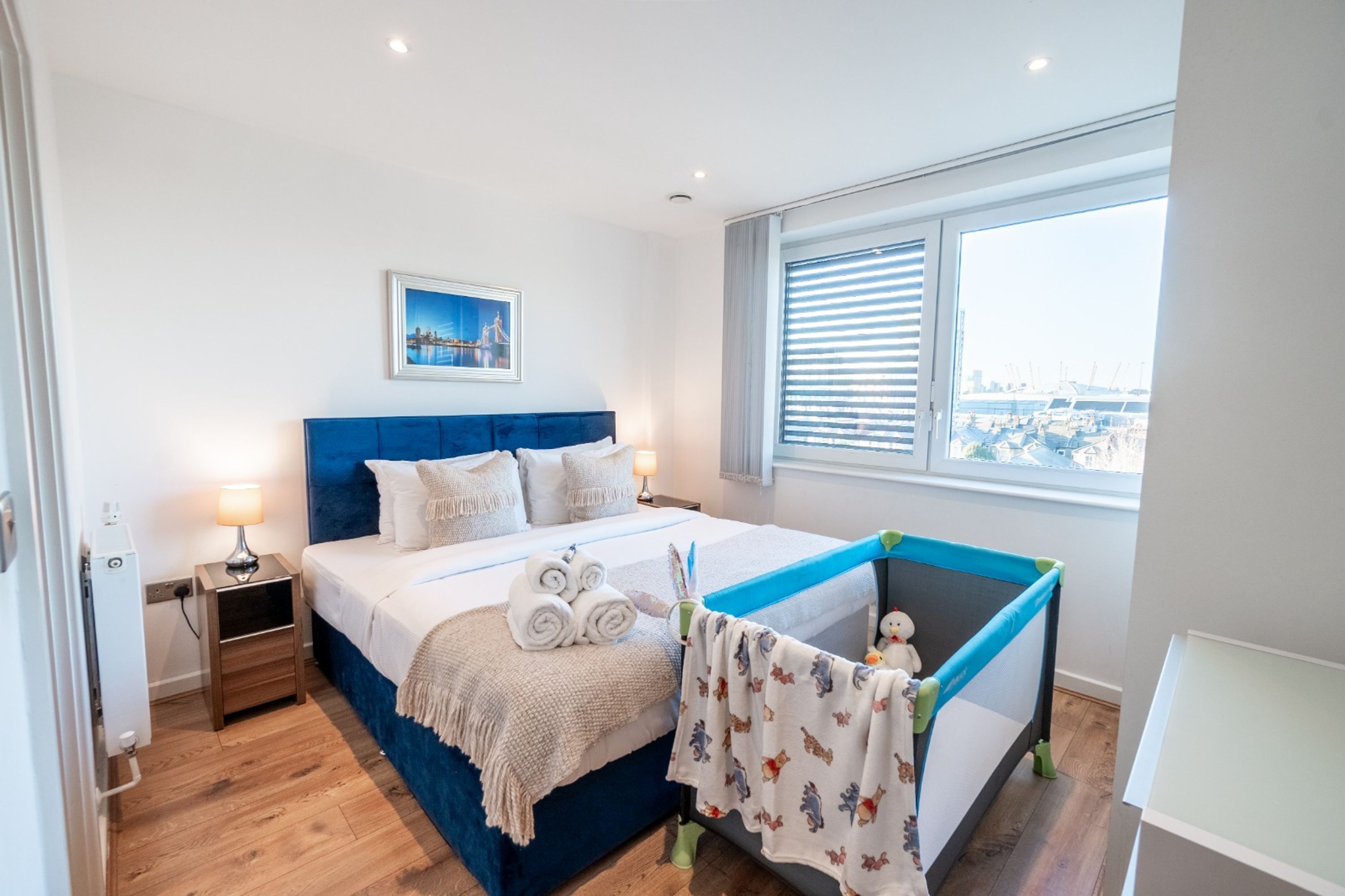 Un dormitorio con cama, mesita de noche, ventana con persianas, vista a la ciudad y un pequeño rincón de juegos con juguete.