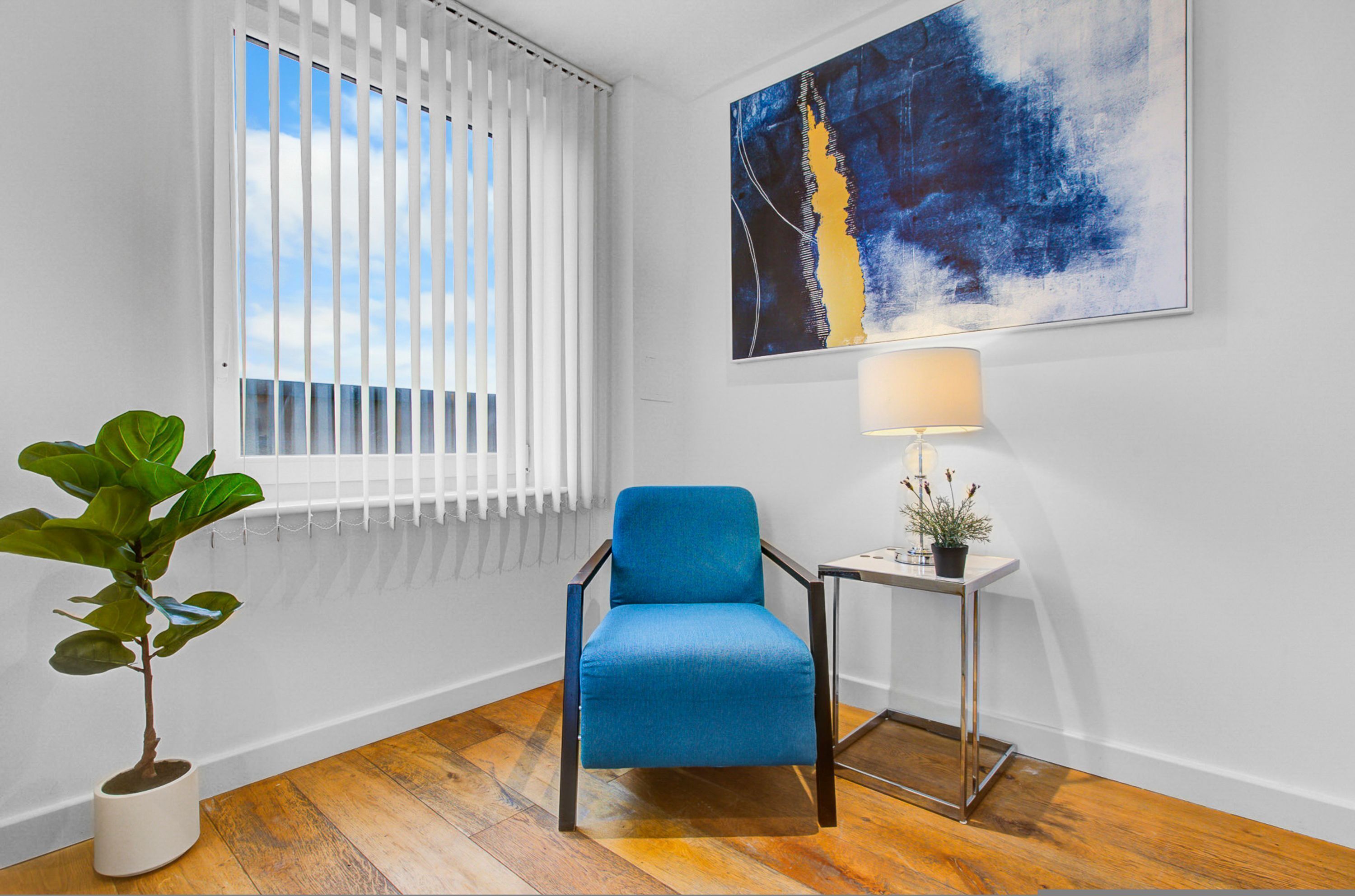 Una habitación con una silla azul, una planta en maceta, una mesa con una lámpara y un cuadro en la pared.
