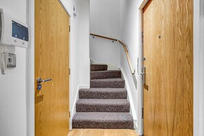 A staircase with carpeted steps leading to a landing, flanked by wooden doors with a security panel on the left. - Hybrid Resi - Greenwich - Two-Bedroom Apartment (London)