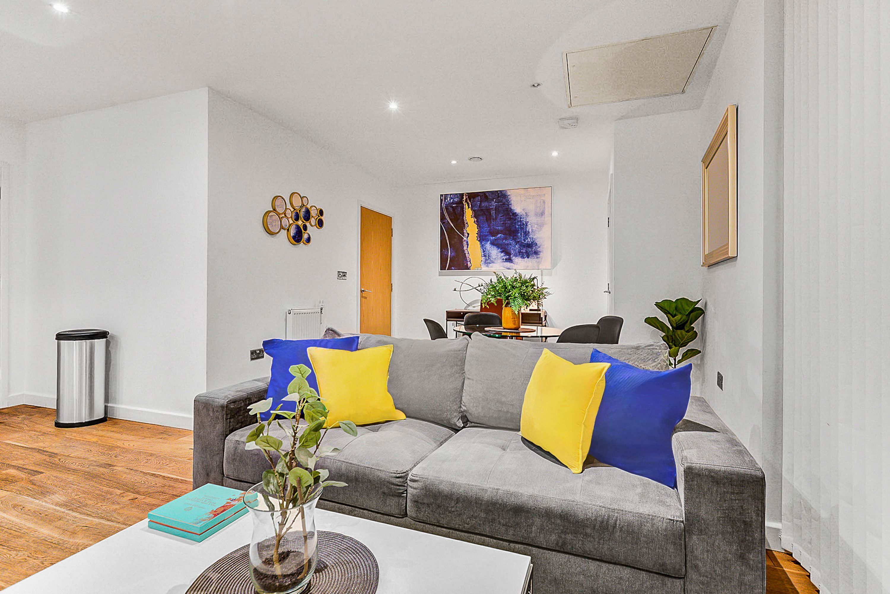 Ein modernes Wohnzimmer mit einem grauen Sofa, gelb-blauen Kissen, einem Couchtisch mit Pflanze und einem Gemälde an der Wand.