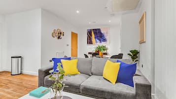 Ein modernes Wohnzimmer mit einem grauen Sofa, gelb-blauen Kissen, einem Couchtisch mit Pflanze und einem Gemälde an der Wand.