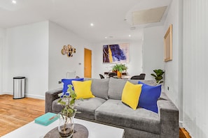 A modern living room with a grey sofa, yellow and blue cushions, a coffee table with a plant, and a painting on the wall. - Hybrid Resi - Greenwich - Two-Bedroom Apartment (London)