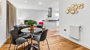 Eine moderne Küche mit Kochinsel, ein Essbereich mit Glastisch und schwarzen Stühlen sowie ein Wohnbereich mit Sofa und Fernseher.