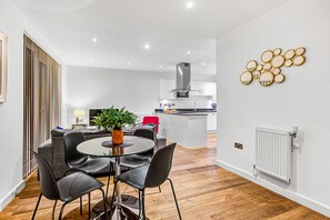 A modern kitchen with a central island, a dining area with a glass table and black chairs, and a living area with a sofa and a TV. - Hybrid Resi - Greenwich - Two-Bedroom Apartment (London)