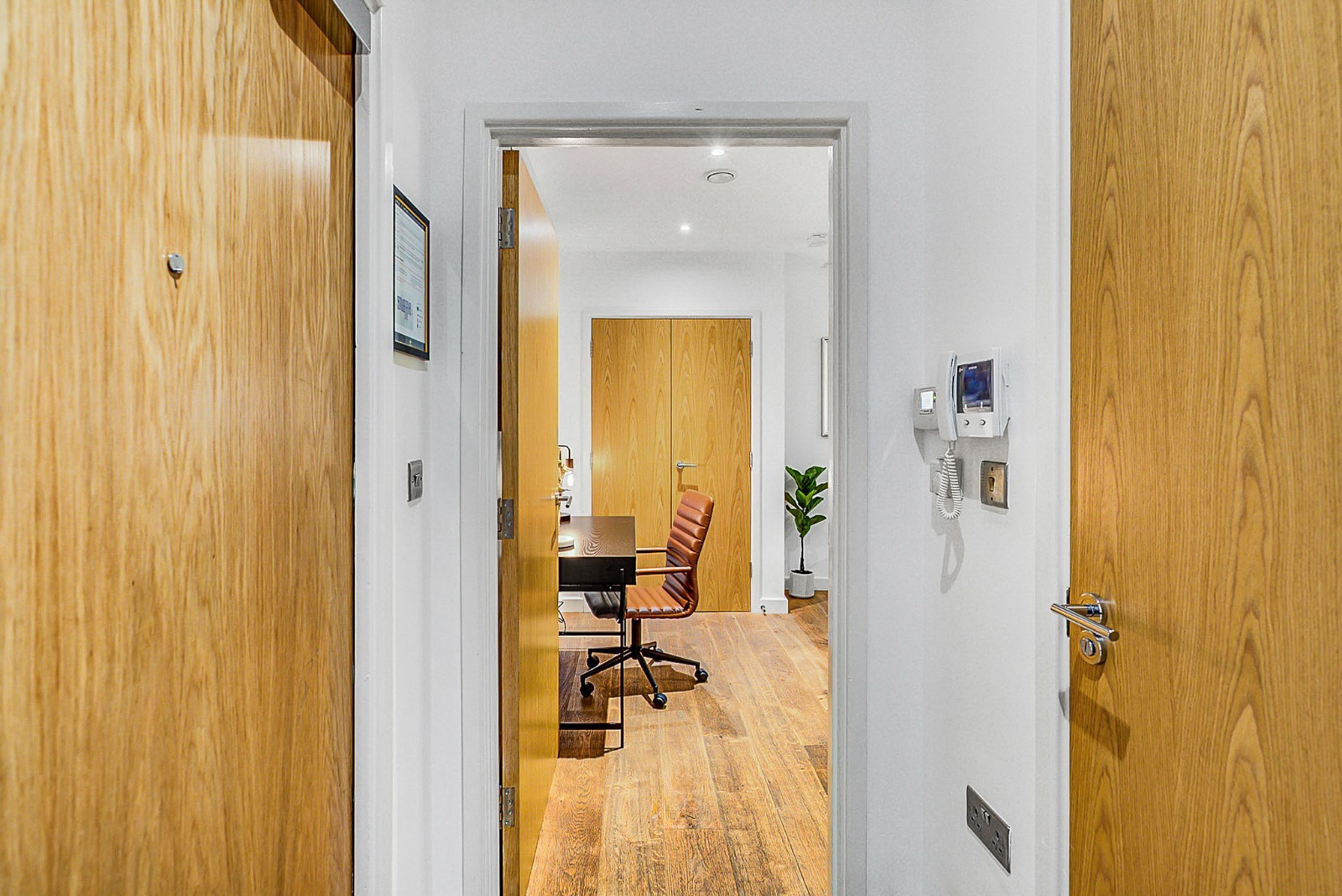A hallway leading to a room with a wooden door, a desk, a chair, and a potted plant.