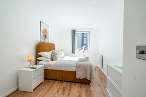 A modern bedroom with a wooden bed, white bedding, a nightstand with a lamp, and a window with a view of the city. - Hybrid Resi - Greenwich - Three Bedroom Apartment (London)