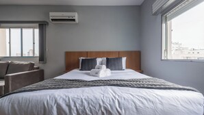 A hotel room with a bed, a window, a chair, and an air conditioning unit.