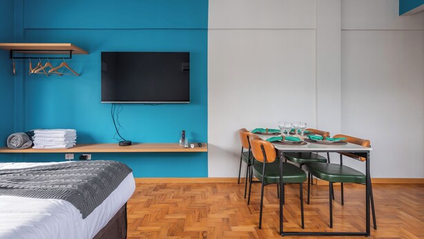 A modern room with a blue wall, a flat-screen TV mounted above a wooden shelf, a bed with a grey blanket, and a dining area with a table set for four. - StayHome Ponta da Praia - 2min da Praia (Santos)