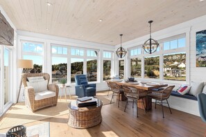 A spacious room with a high ceiling, large windows, and a dining area with wicker furniture.