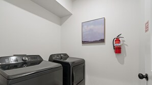 A laundry room with two front-loading washing machines, a fire extinguisher mounted on the wall, and a framed picture above the machines. - Lux Lake View: Sleeps 12+, Hot Tub, Game Room, Theater, Fire Pit (Branson)
