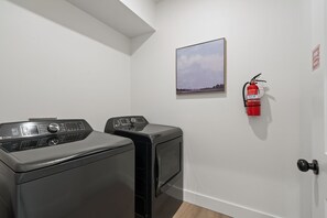 A laundry room with two front-loading washing machines, a fire extinguisher mounted on the wall, and a framed picture above the machines. - Lux Lake View: Sleeps 12+, Hot Tub, Game Room, Theater, Fire Pit (Branson)
