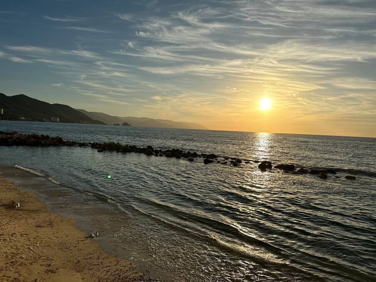 A beach with a rocky breakwater, calm water, and a sunset.