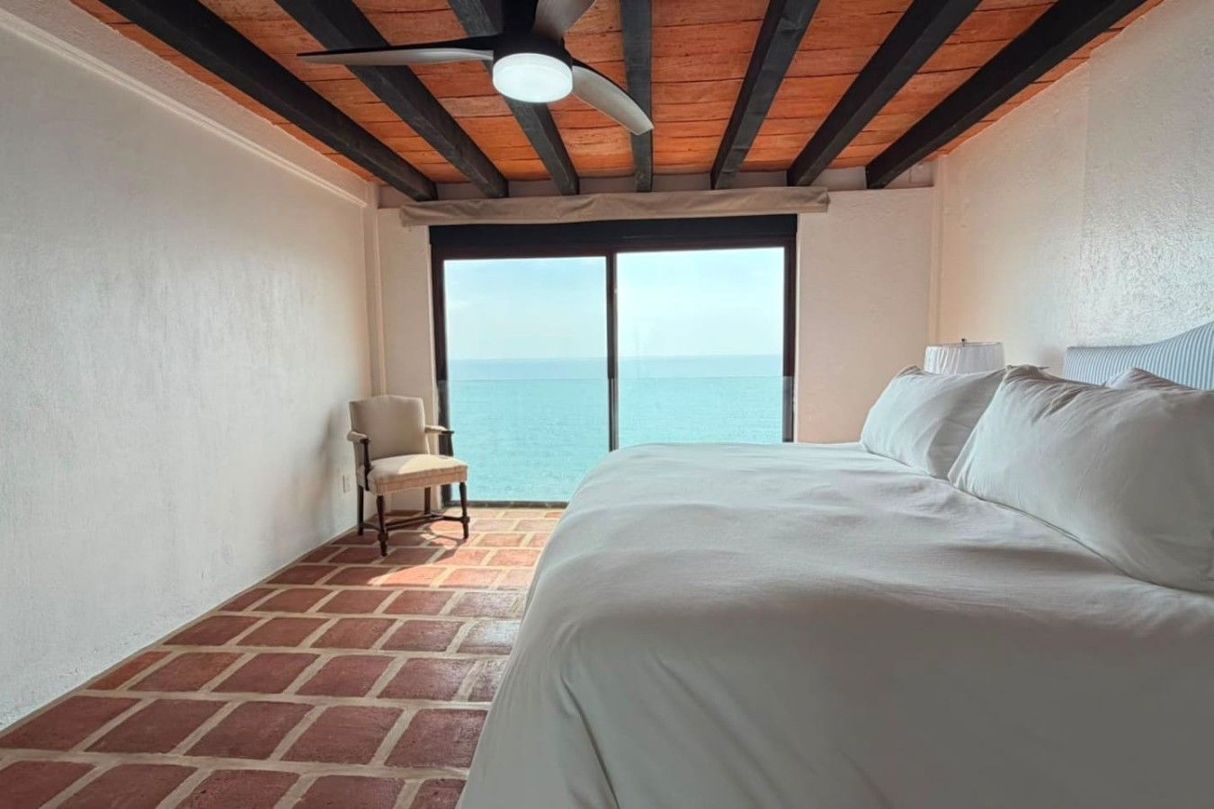 A hotel room with a bed, a chair, a ceiling fan, and a view of the sea through a large window.