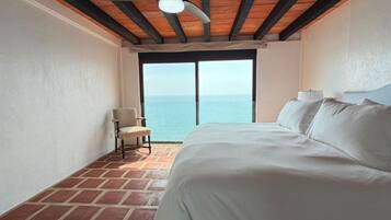 A hotel room with a bed, a chair, a ceiling fan, and a view of the sea through a large window.