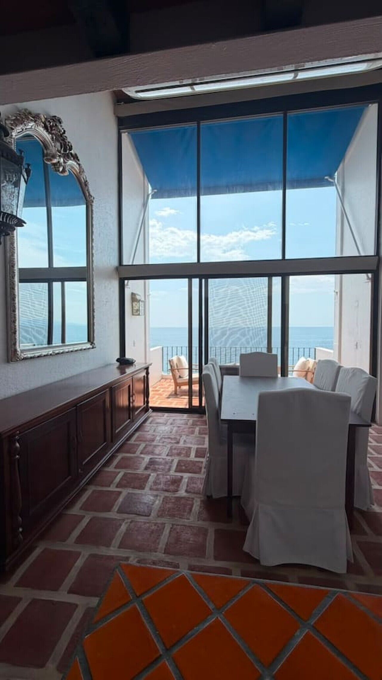 A room with a large window overlooking the sea, a dining area with a table and chairs, and a patterned floor.