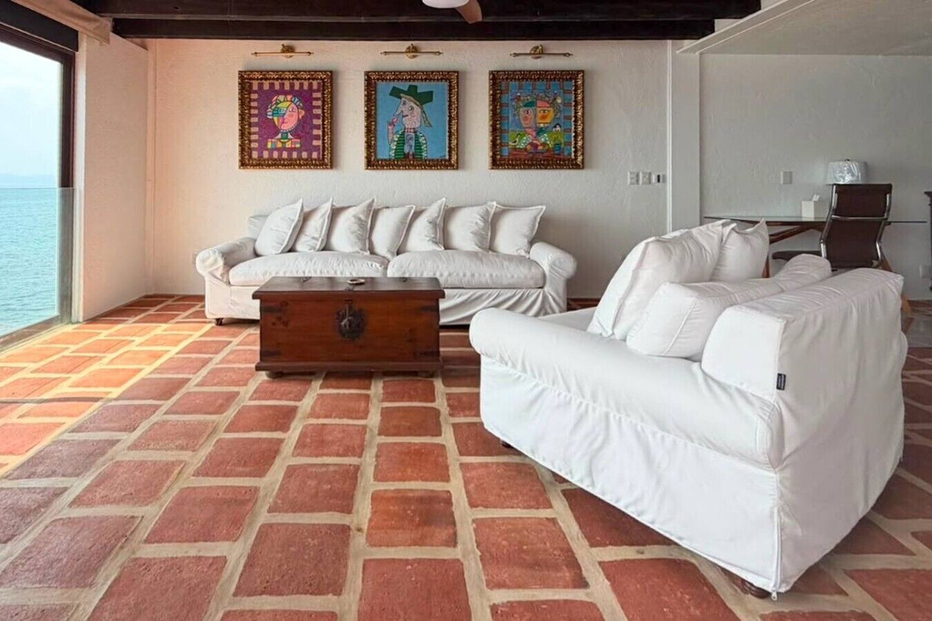 A room with a white sofa set, a wooden chest, and three colorful paintings on the wall.