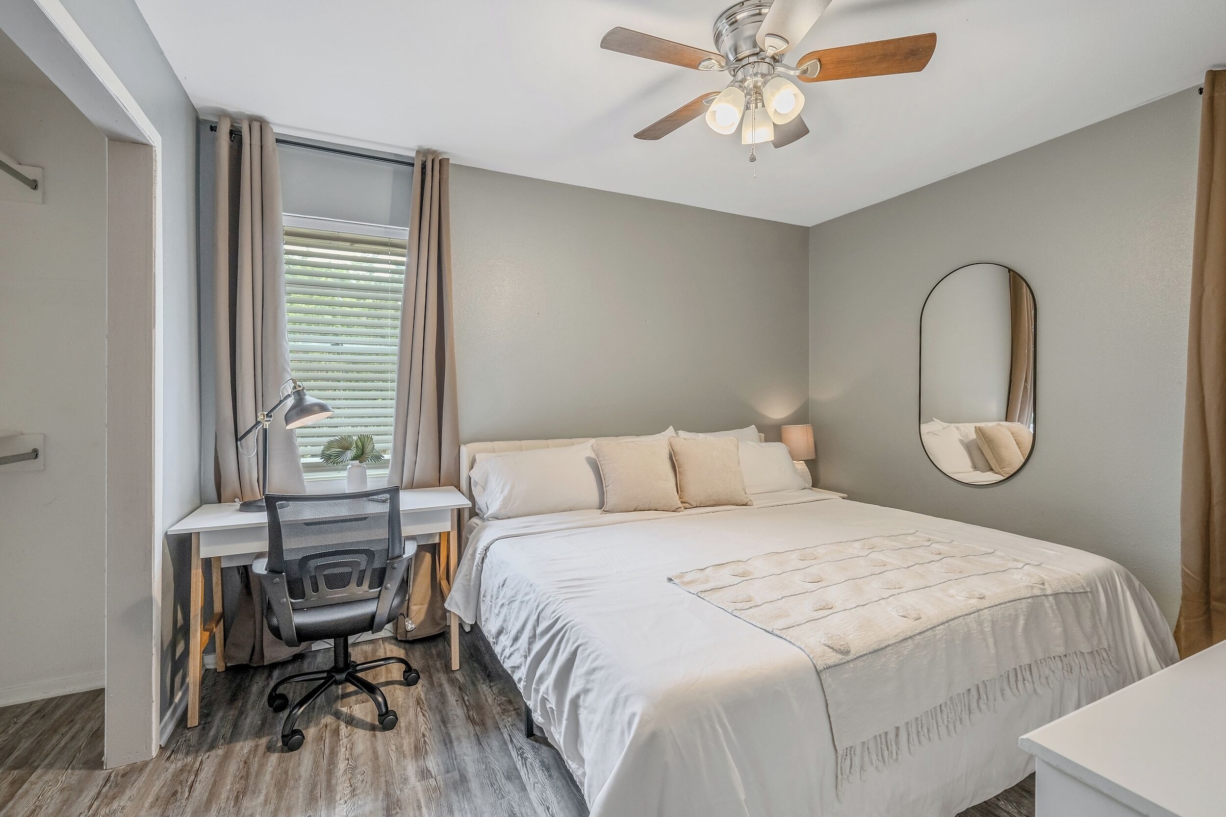 A bedroom with a bed, desk, chair, ceiling fan, window with blinds, and a mirror.