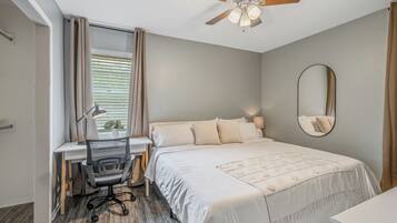 A bedroom with a bed, desk, chair, ceiling fan, window with blinds, and a mirror.