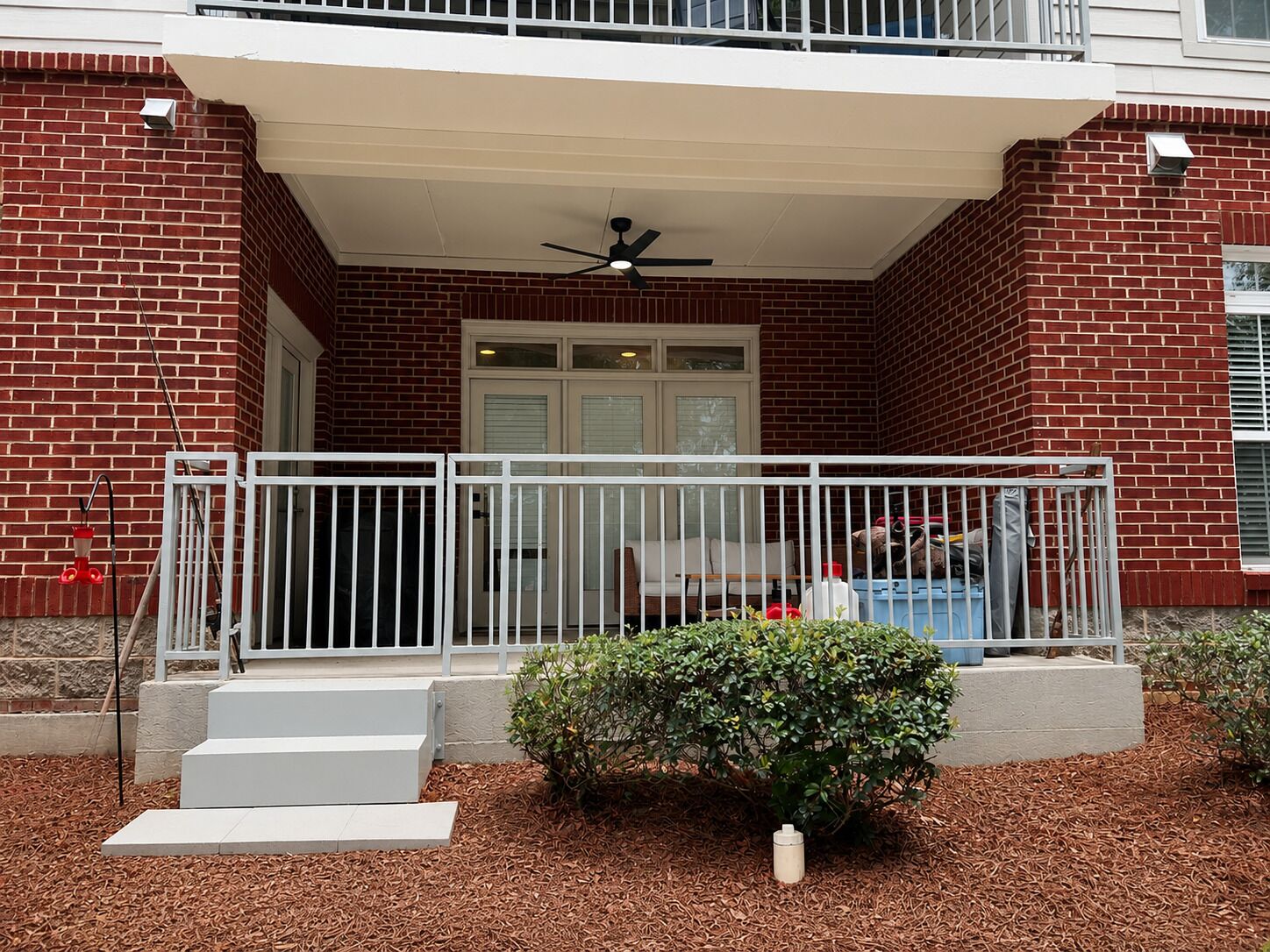 A building with a brick facade, a white balcony railing, and a small garden area with mulch and a bush.