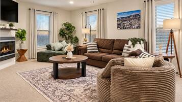 A living room with a fireplace, a ceiling fan, a large window with blinds, and a cityscape painting.