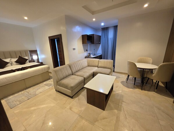 A modern living room with a sectional sofa, a small dining table, and a separate bedroom area with a bed. - Movela Hotels Limited (Warri)