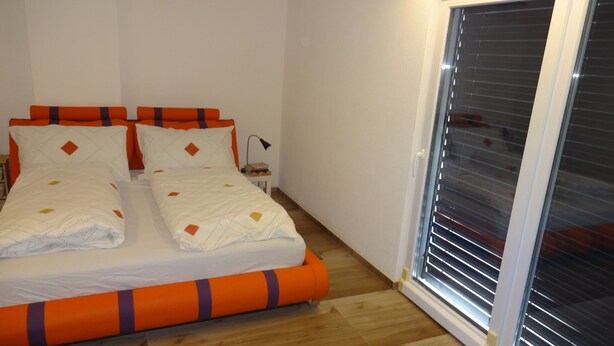 Una cama doble con ropa de cama naranja y blanca, una mesita de noche con lámpara y una puerta corrediza de vidrio a la derecha.