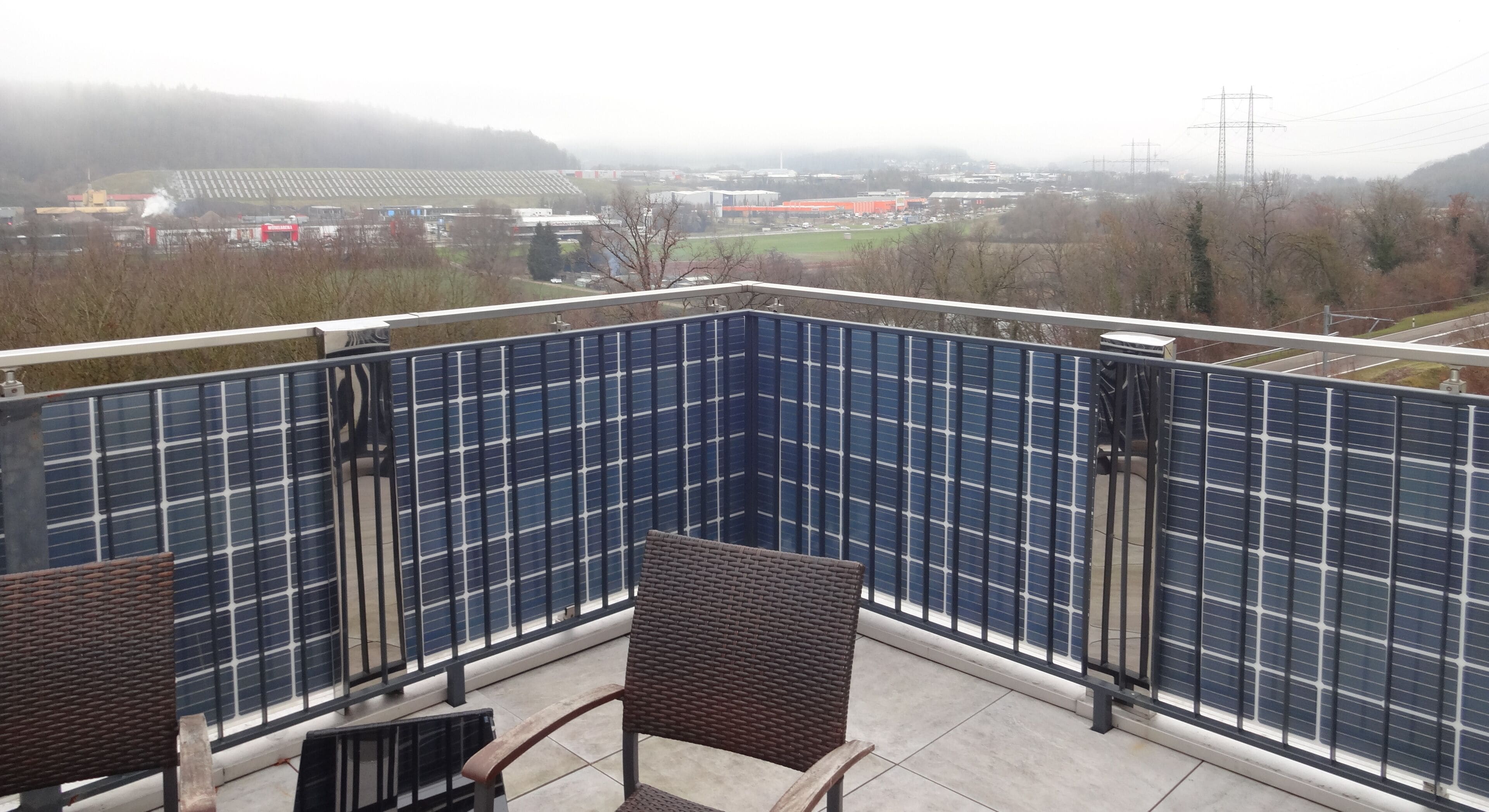 Un balcon agrémenté de panneaux solaires, une chaise en osier et une vue sur une zone industrielle éloignée.