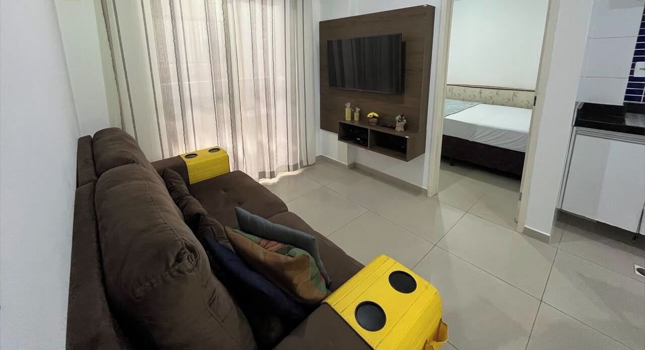 Una sala de estar con un sofá, un televisor montado en la pared, una cama al fondo y una zona de cocina.