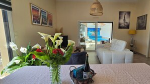 Sala de estar con un sofá blanco, una mesa de comedor con un florero, una botella de champán y vista a una piscina a través de puertas de vidrio.