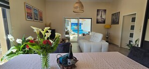 Ein Wohnzimmer mit einem weißen Sofa, einem Esstisch mit Blumenvase, einer Champagnerflasche und Blick auf einen Pool durch Glastüren.