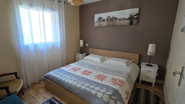 Un dormitorio con una cama, una silla, una mesita de noche, una lámpara, un espejo y una ventana con cortinas.