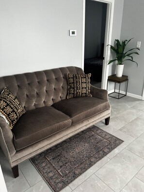 Ein braunes, gebuschtes Sofa mit zwei gemusterten Kissen, ein dunkel gemusterter Teppich und ein Blumentopf auf einem kleinen Tisch daneben.