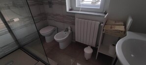 Ein Badezimmer mit Duschkabine aus Glas, zwei Toiletten, einem Bidet, einem Handtuchhalter mit gefalteten Handtüchern und einem weißen Waschbecken.